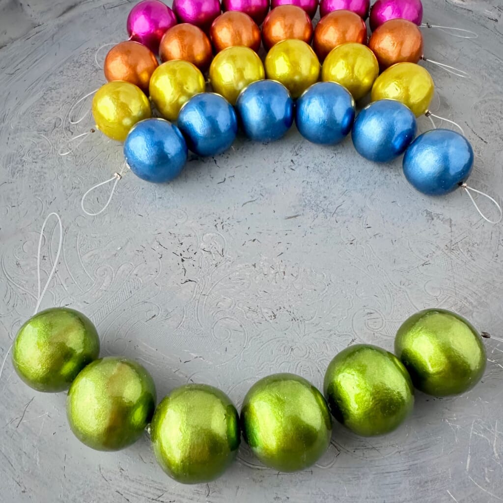 One strand of 6, 24mm round, grass green coloured, cotton pearl beads with a pearlized, lightweight, textured finish, shown on an embossed grey background, from Suzie Q Studio.