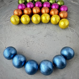 One strand of 6, 24mm round, Montana blue coloured, cotton pearl beads with a pearlized, lightweight, textured finish, shown on an embossed grey background, from Suzie Q Studio.