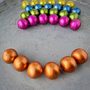 One strand of 6, 24mm round, orange coloured, cotton pearl beads with a pearlized, lightweight and textured finish, shown on an embossed grey background, from Suzie Q Studio.