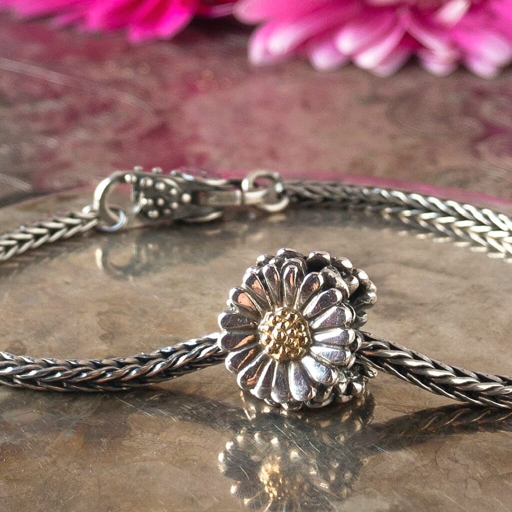 Trollbeads sterling silver & 18 karat gold DAISY bead, shown on a sterling silver Trollbeads bracelet, with pink daisy flowers in background.