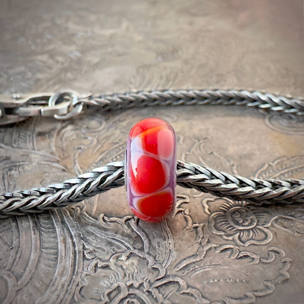Available at Suzie Q Studio dot com, Trollbeads retired LILAC EDGE, glass bead, with red, "stepping  stone" shapes, floating on a warm lilac-purple base, shown on a silver chain with a clasp.