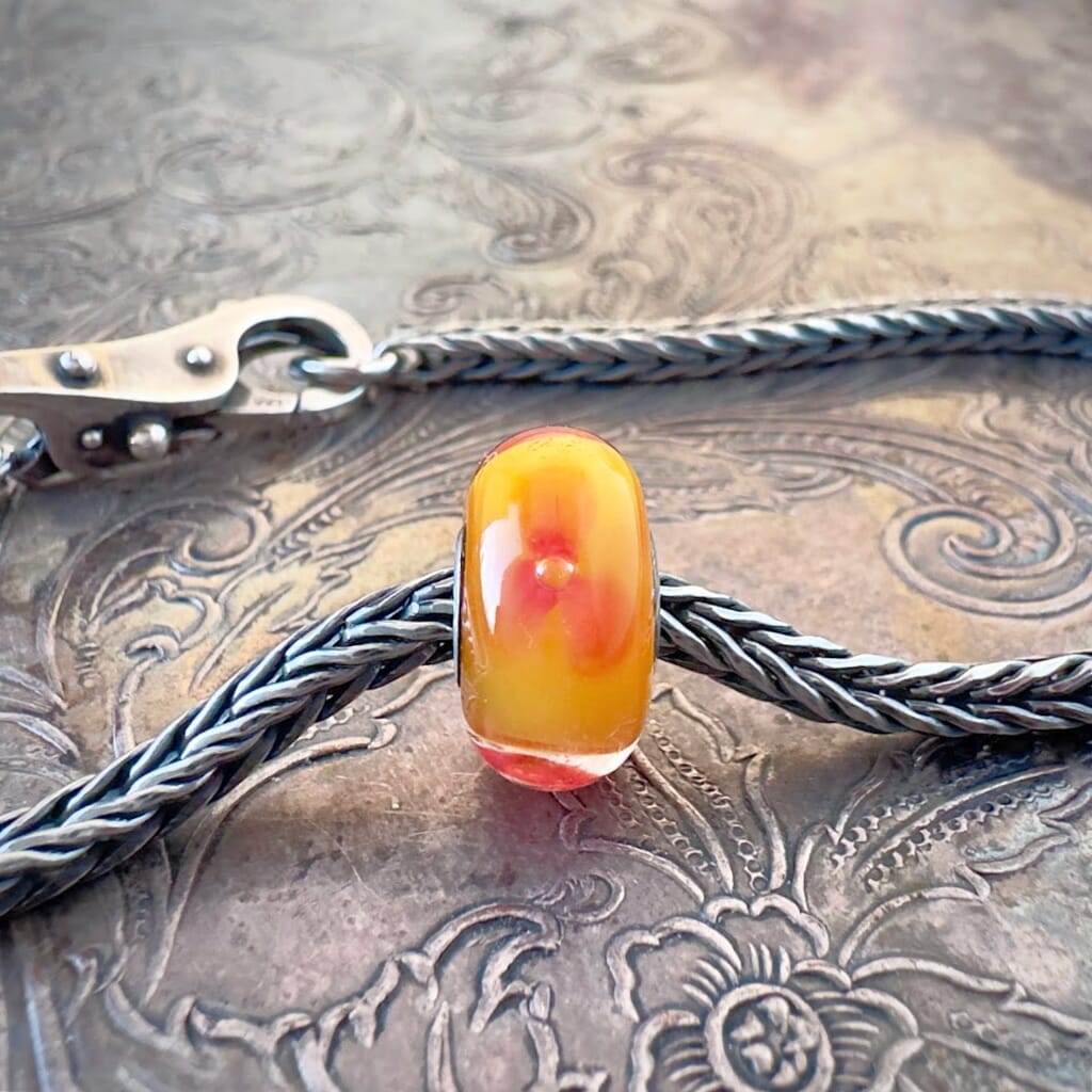Available at Suzie Q Studio dot com, Trollbeads retired RED FLOWER, glass bead, with a trio of red flowers floating on a warm orange base, shown on a silver chain with a clasp.