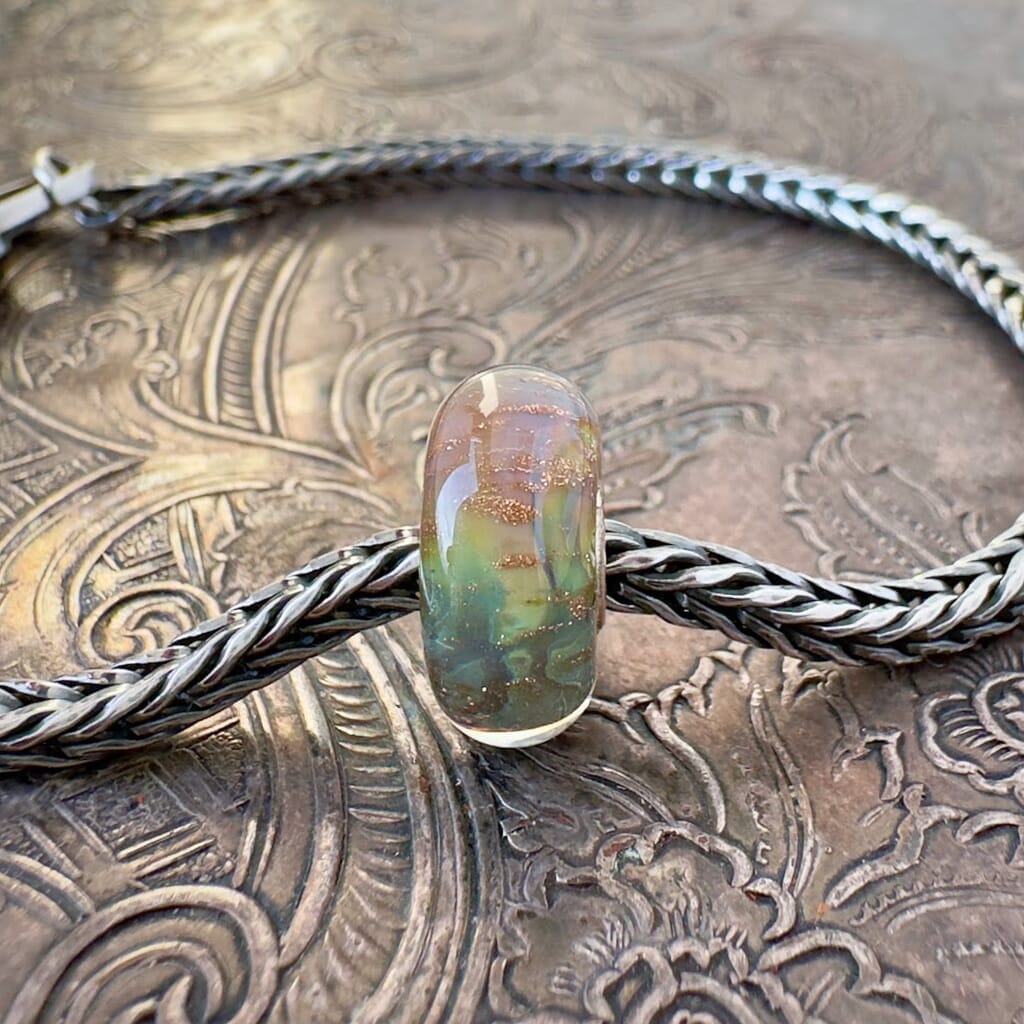 Trollbeads, retired, SHADES OF PEACE, glass bead, featuring earthy, blue-green-ochre colours and copper glitter, which is shown on a sterling silver Trollbeads bracelet, with a clasp, on an embossed background, and is available at Suzie Q Studio dot com.