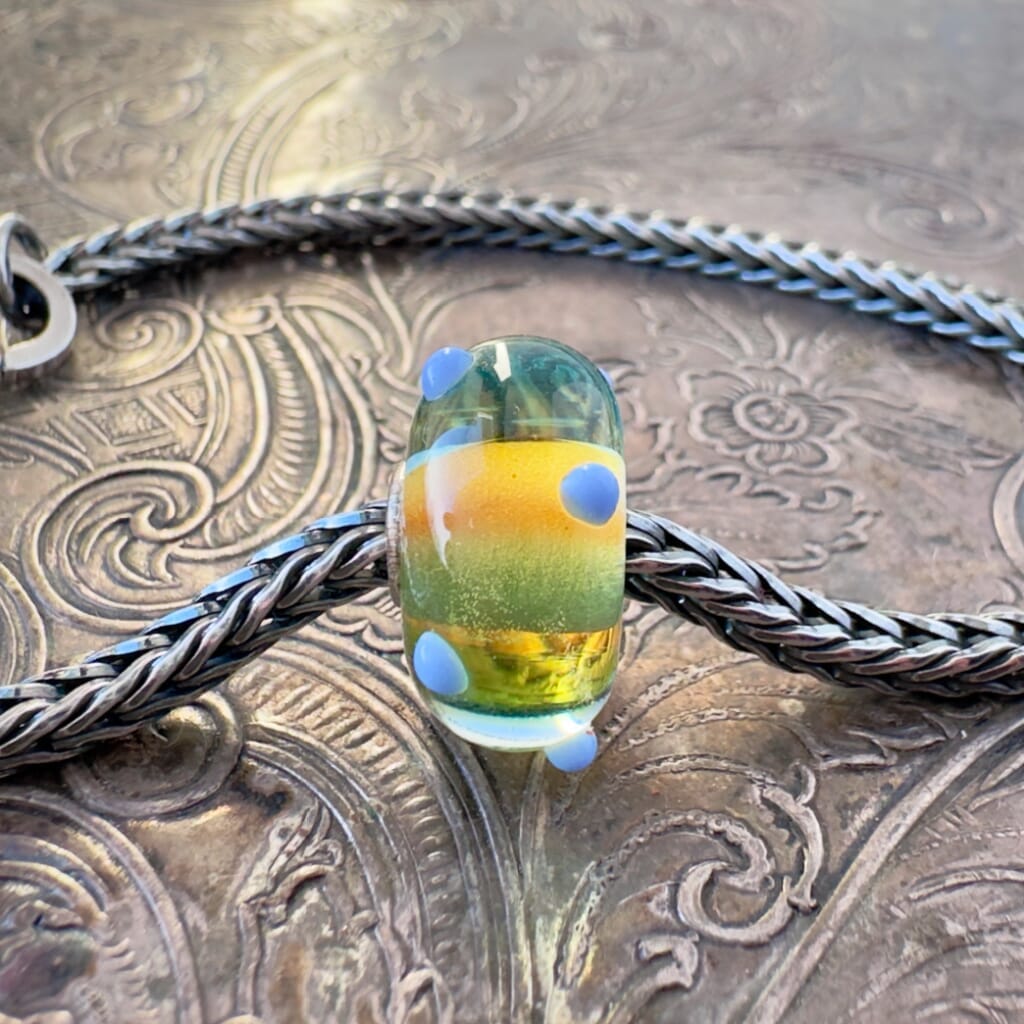 Available at Suzie Q Studio dot com, this Trollbeads, jumbo-sized, UNIQUES, "RAINBOW-BUD" style, glass bead, features a rainbow of colours in the interior of the bead, which is encased in a super-thick coat of clear glass, along with 3-D,  opaque, pale blue buds around the  outside. This fabulous bead is shown on a silver Trollbeads chain with a clasp, on an embossed background.