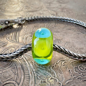 Available at Suzie Q Studio dot com, this Trollbeads, jumbo-sized, UNIQUES, "BUBBLES" style glass bead, features large, alternating circles of opaque lime green and turquoise, with a bubble embedded in the centre of each one, which are floating on a base of transparent lime green. This fabulous bead is shown on a silver Trollbeads chain with a clasp, on an embossed background.