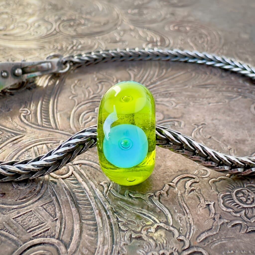 Available at Suzie Q Studio dot com, this Trollbeads, jumbo-sized, UNIQUES, "BUBBLES" style glass bead, features large, alternating circles of opaque lime green and turquoise, with a bubble embedded in the centre of each one, which are floating on a base of transparent lime green. This fabulous bead is shown on a silver Trollbeads chain with a clasp, on an embossed background.