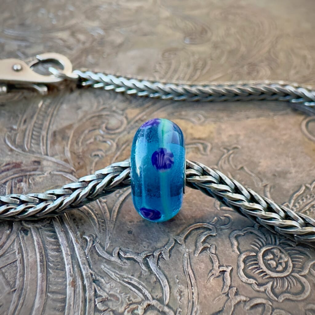 Available at Suzie Q Studio dot com, Trollbeads retired CHINA BLUE, glass bead, with strands of soft green and dark blue circles, floating on a clear, vibrant blue base, shown on a silver chain with a clasp.