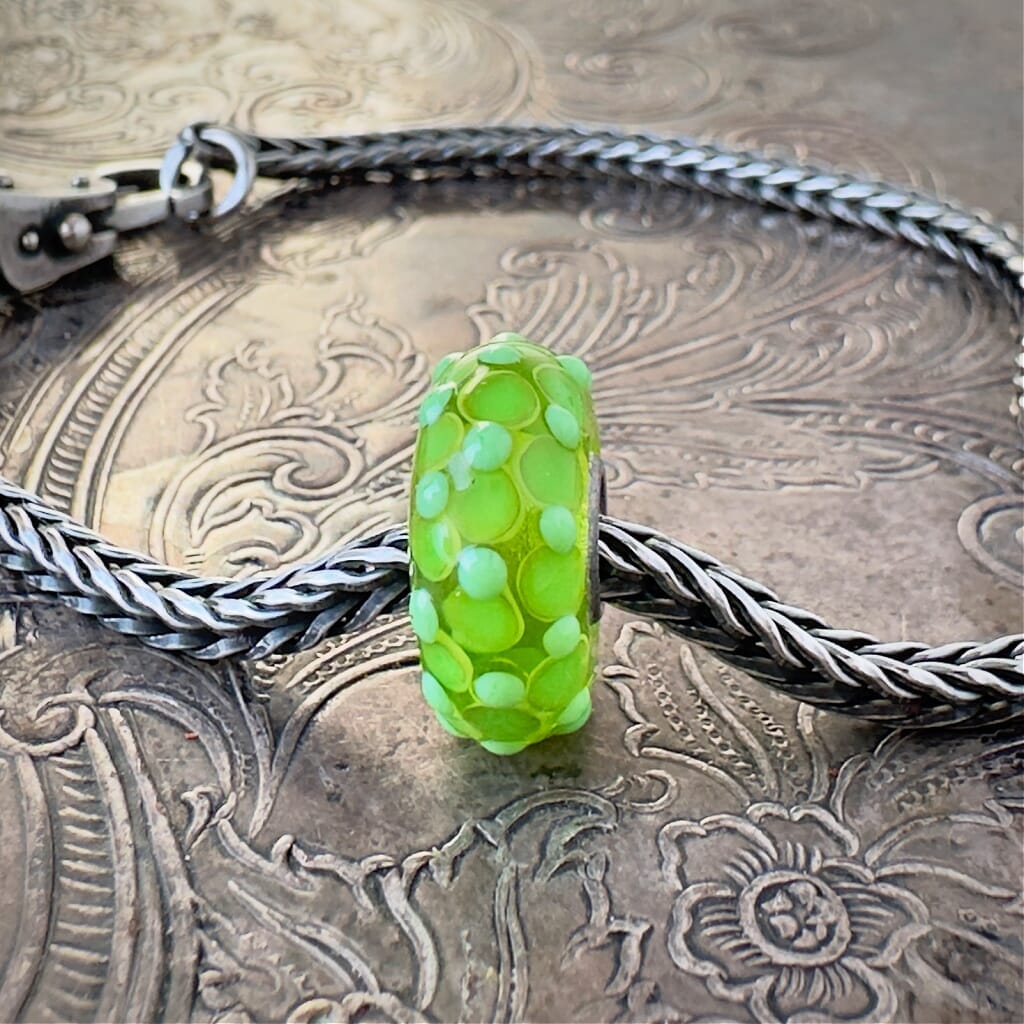 Available at Suzie Q Studio dot com, Trollbeads UNIVERSAL UNIQUES, one-of-a-kind, transparent, lime green, glass bead, embellished with dots and delicate, 3-D, textural bumps, in various tones of lime green, and shown on a silver chain with a clasp, on an embossed background.