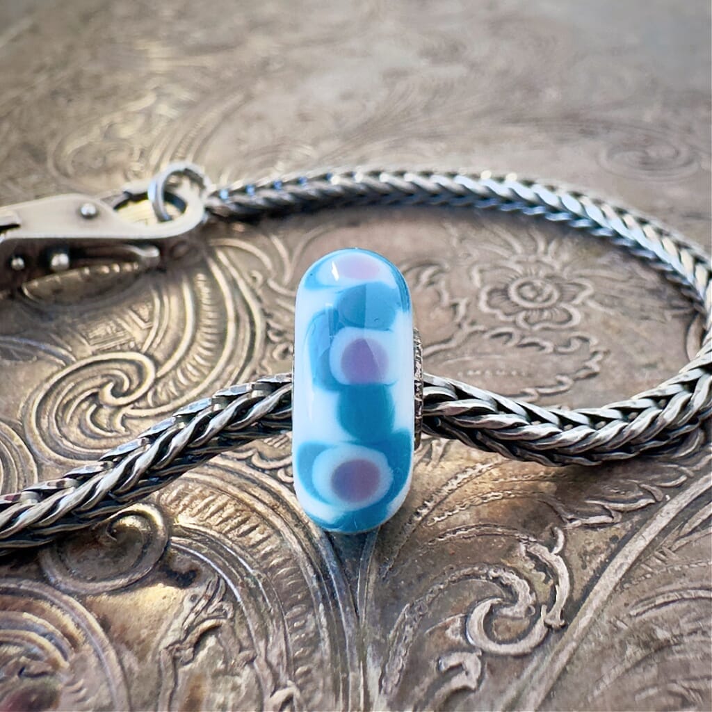 Available at Suzie Q Studio dot com, Trollbeads UNIVERSAL UNIQUES, one-of-a-kind, glass bead, is what's known in Troll World as a "Big Dimples Smile" design, in opaque, medium blue and lilac, on a base of opaque white, and is shown on a Trollbeads silver chain, with a clasp, on an embossed background.