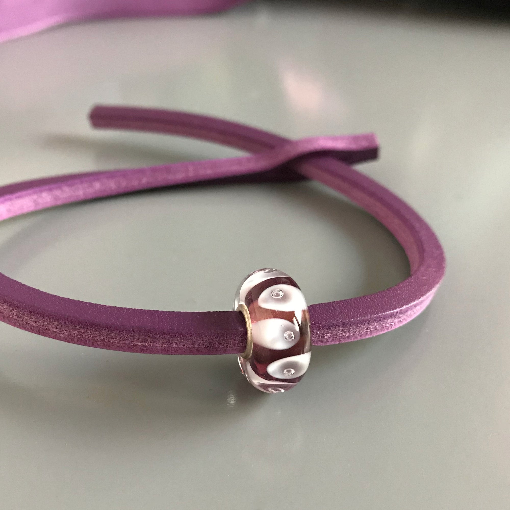 Trollbeads UNIQUES are one-of-a-kind glass beads hand made individually by 100% artisan-owned workshops in Tibet, India, Malawi and Lithuania. This Suzie Q Studio UNIQUES glass bead has white bubbles dancing on a transparent amethyst-coloured background and includesa leather bracelet.